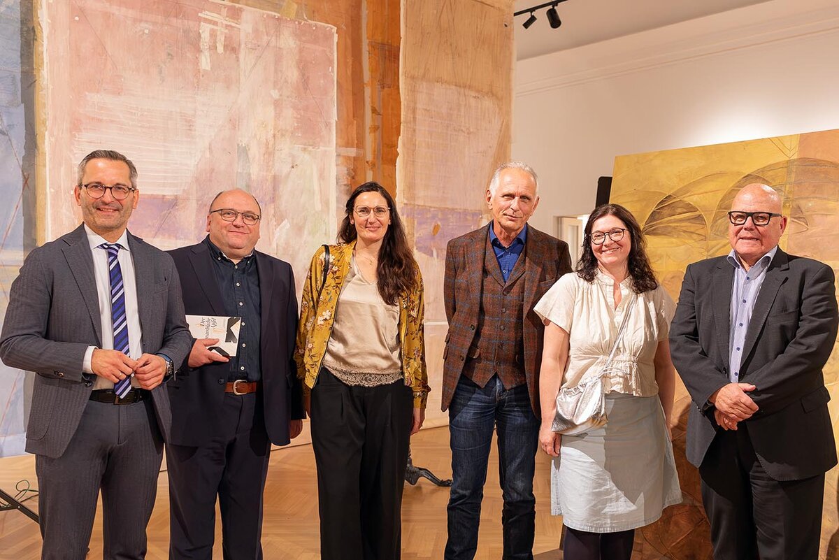 Sechs Personen vor Kunstwerken, darunter ein Mann mit Buch in der Hand.