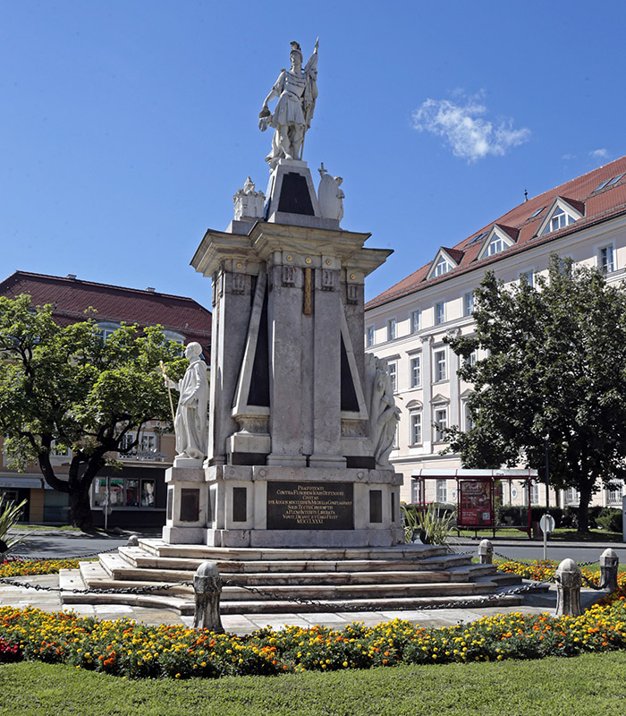 Florianidenkmal mit Grün und Blumenbepflazung rundherum, im Hintergrund das Gerichtsgebäude©StadtPresse/Eggenberger
