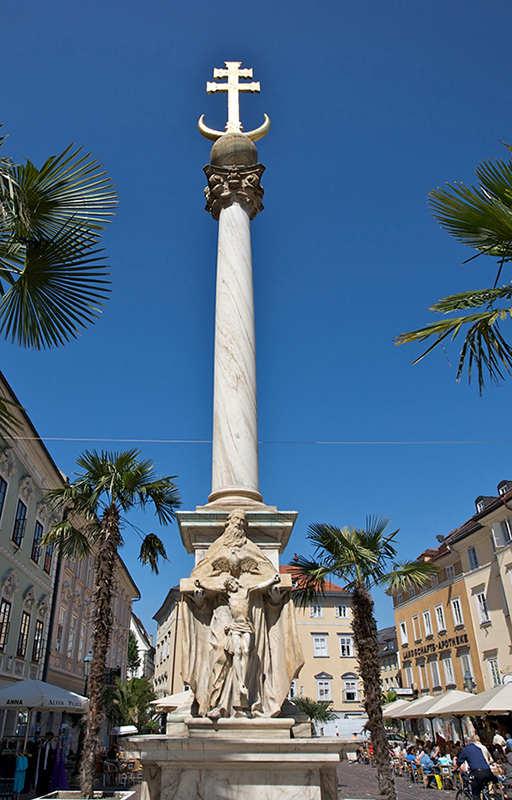 Hoch ragt die Pestsäule auf dem Alten Platz gegen Himmel, umgeben von Palmen©StadtPresse