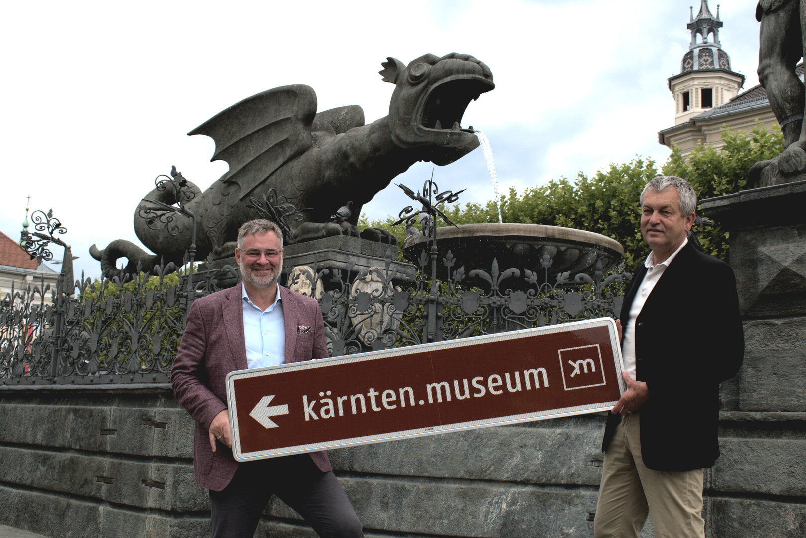 Adi Kulterer, Vorstand Tourismusverband Klagenfurt am Wörthersee, und Tourismusstadtrat Max Habenicht mit einem Beispielschild vorm Lindwurm. Foto: Büro Habenicht